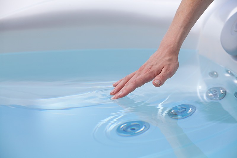 A hand reaching into the blue water of a hot tub, creating ripples. Keeping hot tub water clean is important to prevent algae and mildew growth.